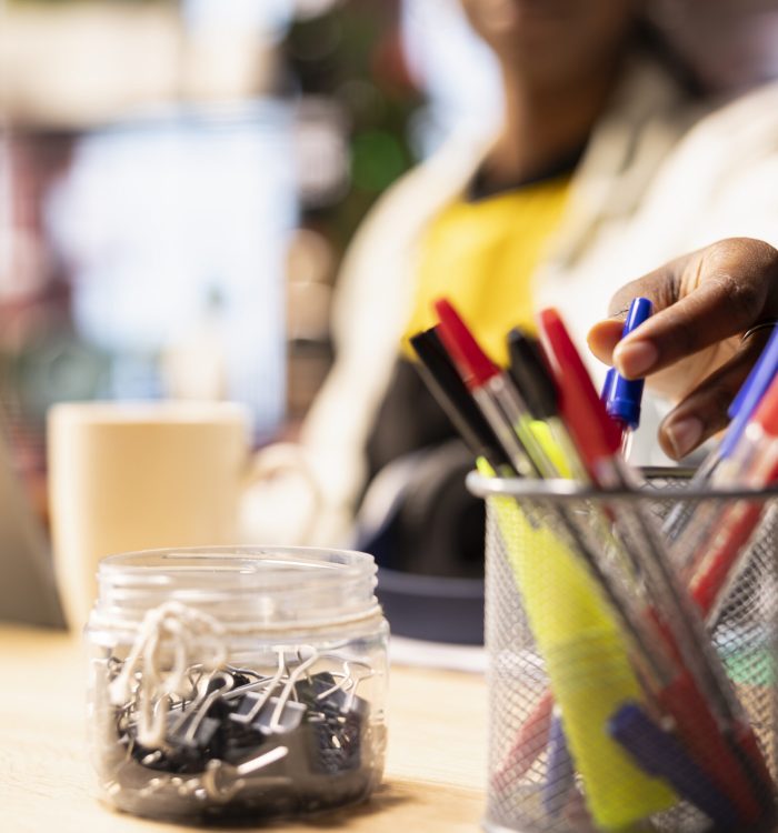 Close up of student in home office reaching for pen from desk organizer, preparing to jot down important notes. Focus on workspace stationary holder used by person in blurry background to do writing